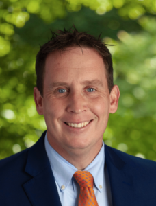 Ian Moorhouse, a white man with short brown hair and blue eyes, wears a blue suit and orange tie against a green background of trees. He is smiling.